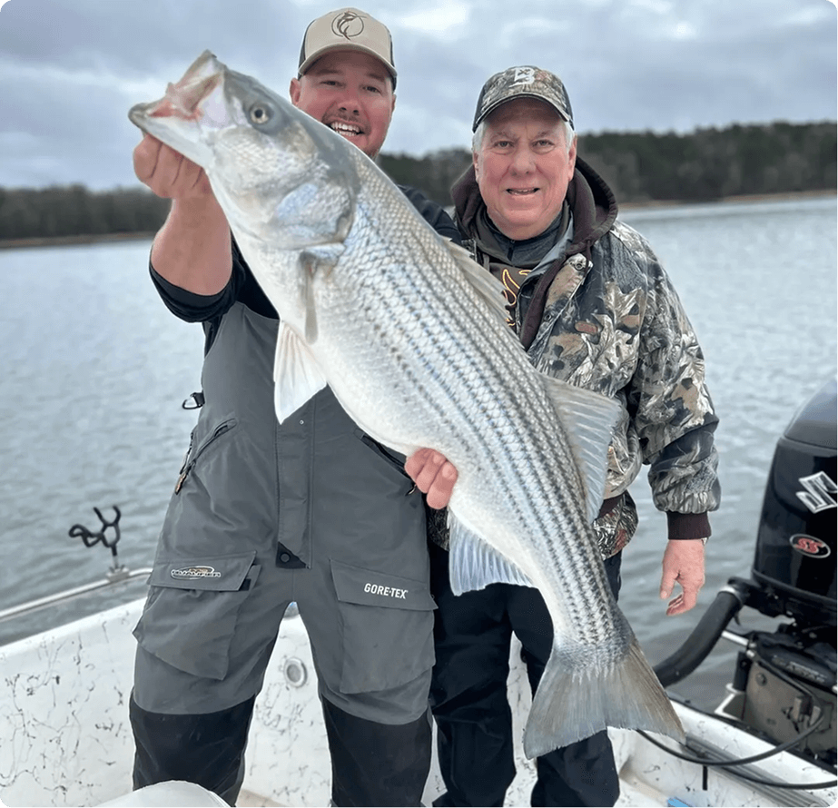 Two men on a boat proudly hold a large striped bass. They are dressed in outdoor gear, and the water and trees are visible in the background.